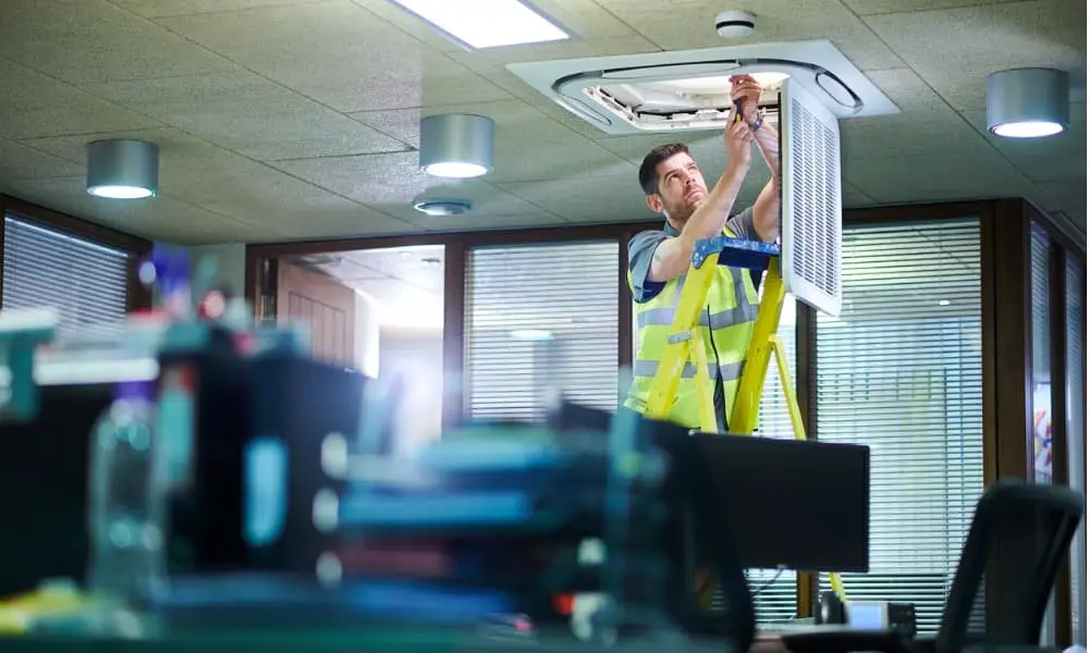Facility maintenance technician on a ladder servicing a ceiling HVAC unit in an office